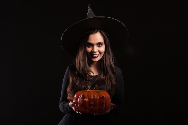 Jonge heks met een grote hoed die een enge pumpking vasthoudt. Mooie jonge vrouw in een halloween-kostuum. Fancy kostuum voor Halloween.