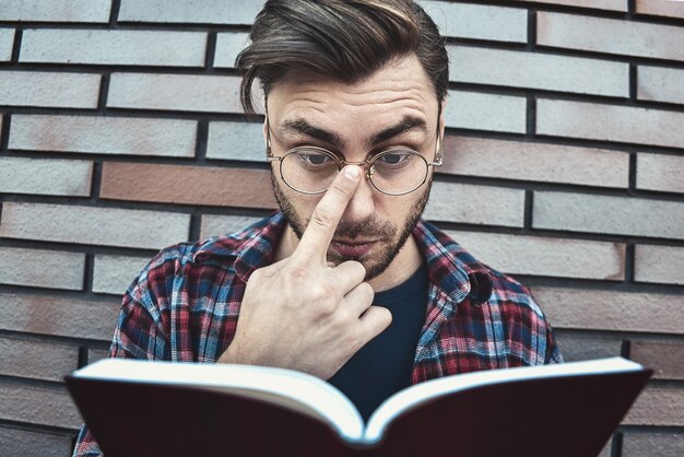 Jonge hipster kerel die een bril draagt die een boek of notitieboek op bakstenen muuroppervlak leest