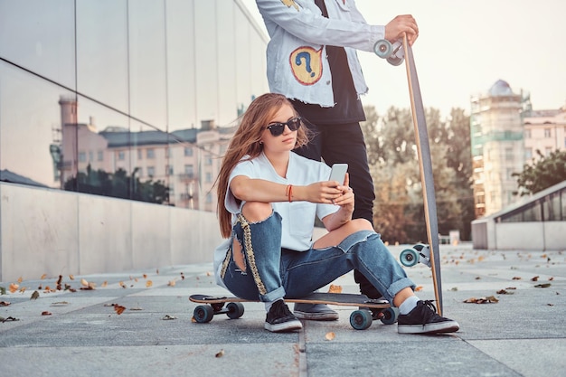 Jonge hipster meisje zittend op een longboard en met behulp van een smartphone terwijl haar vriendje in de buurt staat.