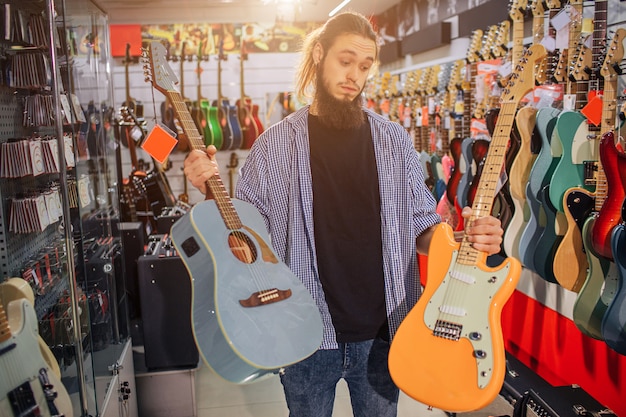 Jonge hipster staat en houdt zowel elektrische als akoestische gitaren vast. Hij kijkt naar de gele. Guy kan niet kiezen tussen twee instrumenten.