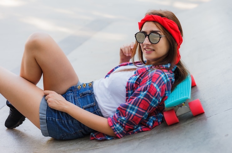 Foto jonge hipster vrouw in stijlvolle spiegelglazen, korte broek, rood geruite overhemd en verband op haar hoofd ligt met skateboard op betonnen ondergrond in skatepark