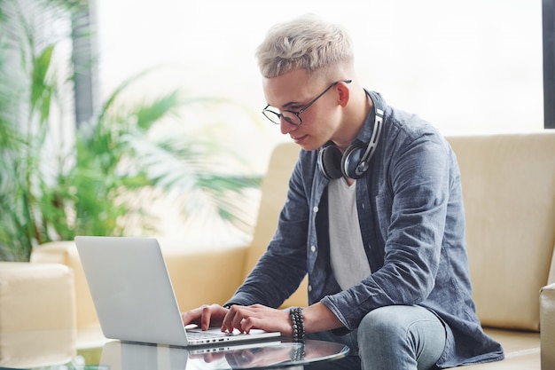 Jonge hipsterkerel die in aardige kleren op bank met laptop zitten