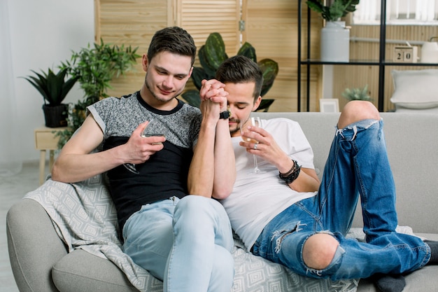 Jonge homoseksuelen die handen op bank thuis houden. Verlegen homopaar dat liefde en romantiek toont, wijn drinkt