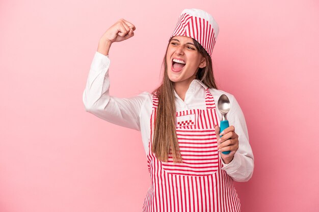 Jonge ijsmachine vrouw met lepel geïsoleerd op roze achtergrond die vuist opheft na een overwinning, winnaar concept.