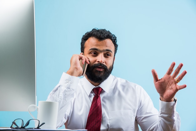 Jonge indiase aziatische zakenman in baard bezig met telefoongesprek tijdens het gebruik van laptop of computer op kantoor