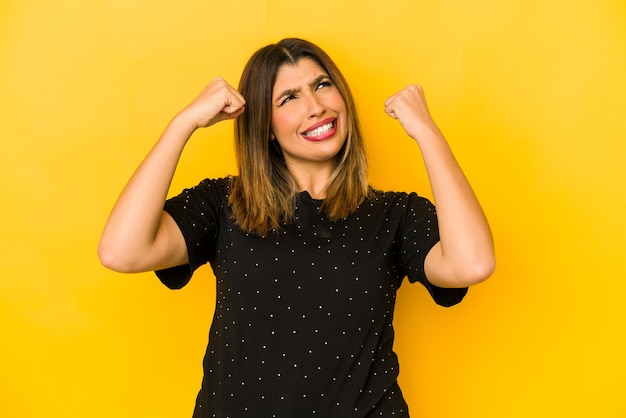 Jonge Indiase die vrouw op gele achtergrond wordt geïsoleerd die vuist opheft na een overwinning, winnaarconcept.