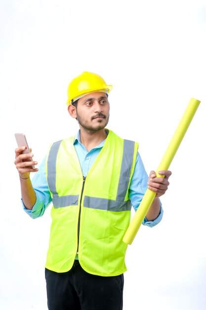 Jonge Indiase ingenieur die veiligheidshelm draagt en smartphone op witte achtergrond gebruikt.