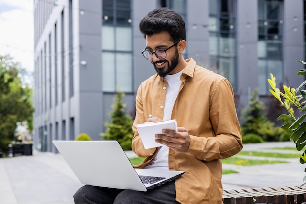 Jonge indiase man die online studeert zittend op een bankje buiten de universiteitscampus student met laptop