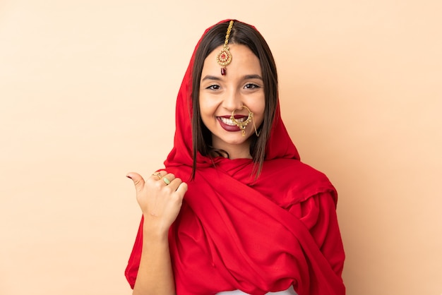 Jonge Indiase vrouw geïsoleerd op beige muur wijzend naar de kant om een product te presenteren