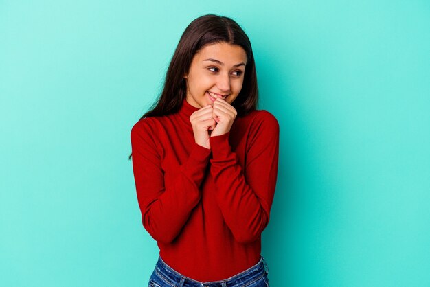 Jonge Indiase vrouw geïsoleerd op blauw houdt handen onder de kin, kijkt gelukkig opzij.