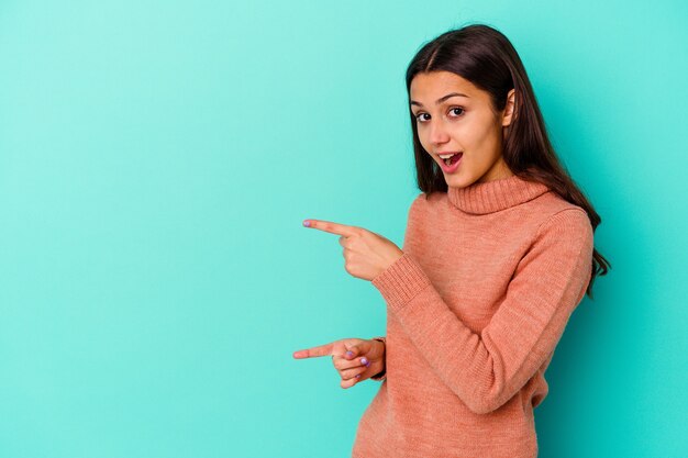 Jonge Indiase vrouw geïsoleerd op blauw opgewonden wijzend met wijsvingers weg.