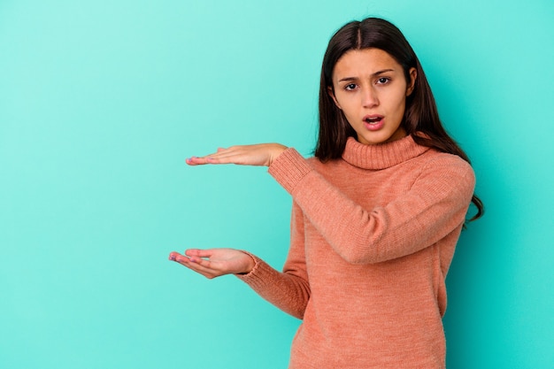 Jonge Indiase vrouw geïsoleerd op blauwe muur geschokt en verbaasd met een kopie ruimte tussen handen.