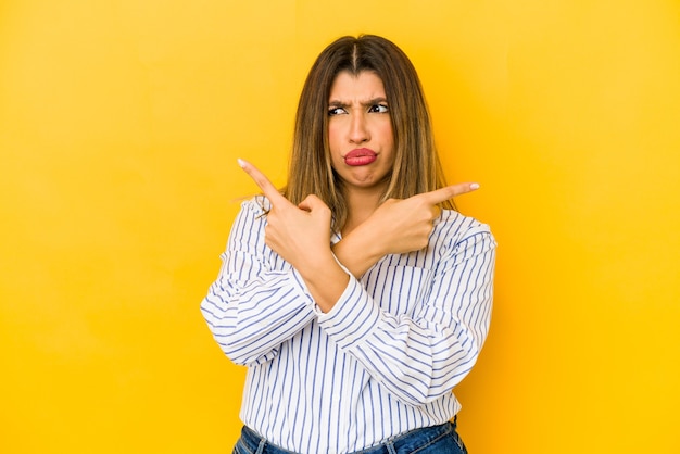 Jonge indiase vrouw geïsoleerd op gele achtergrond punten zijwaarts, probeert te kiezen tussen twee opties.