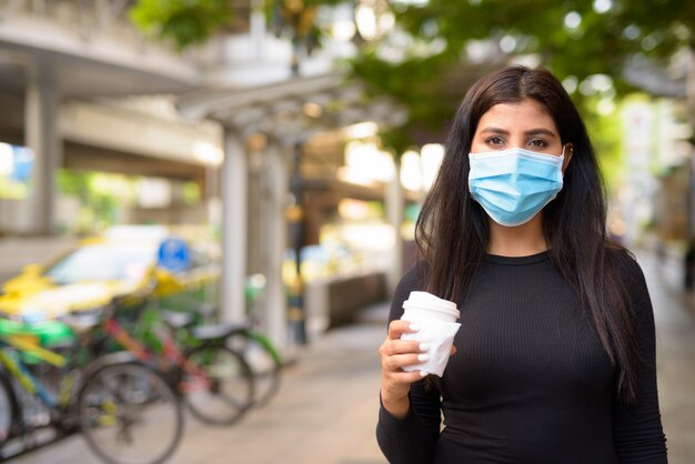 Jonge Indiase vrouw met masker die onderweg koffie drinkt als het nieuwe normaal tijdens covid-19 in de stad