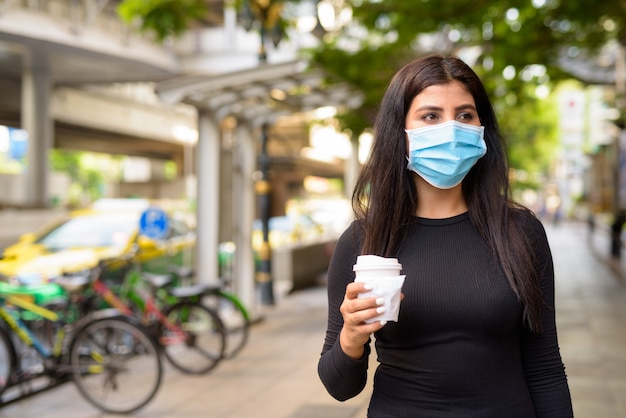 Jonge Indiase vrouw met masker die onderweg koffie drinkt als het nieuwe normaal tijdens covid-19 in de stad