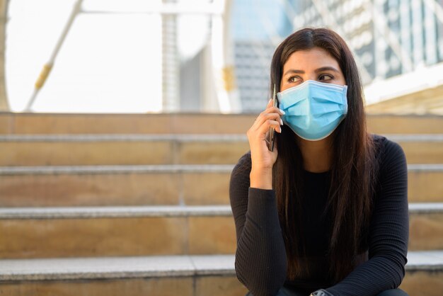 Jonge Indiase vrouw met masker praten aan de telefoon en zitten bij de trap in de stad