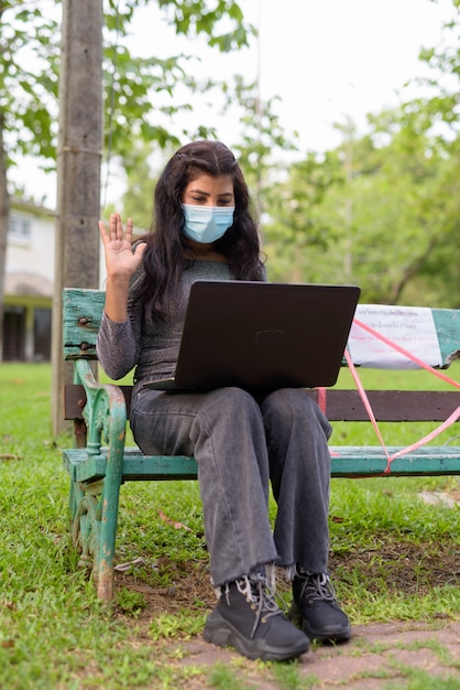 Jonge indiase vrouw met masker videobellen zittend met afstand op bankje