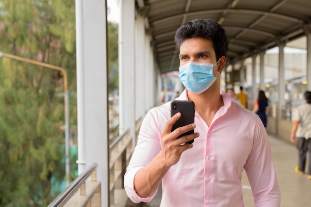 Jonge Indiase zakenman met masker denken tijdens het gebruik van telefoon bij de loopbrug