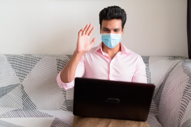 Jonge Indiase zakenman met masker videobellen met behulp van laptop en thuis blijven onder quarantaine