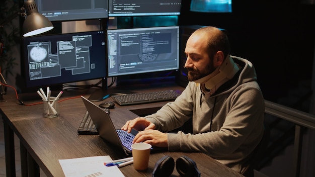 Jonge IT-programmeur draagt halsband op kantoor en typt 's avonds laat servercode op meerdere schermen. App-ontwikkelaar met medische nekbrace die werkt met kunstmatige intelligentie. Schot uit de hand.
