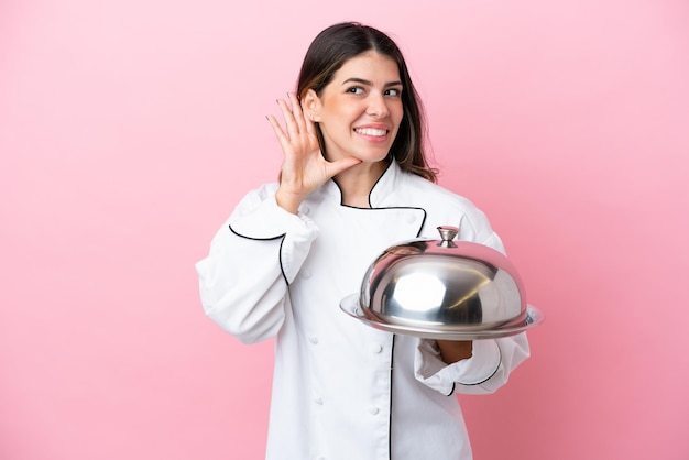 Jonge italiaanse chef-kok vrouw met dienblad met deksel geïsoleerd op roze achtergrond luisteren naar iets door hand op het oor te leggen