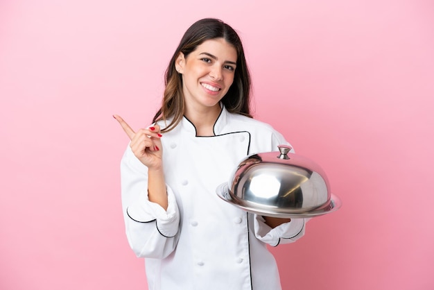 Jonge Italiaanse chef-kokvrouw met dienblad met deksel geïsoleerd op een roze achtergrond die naar de zijkant wijst om een product te presenteren