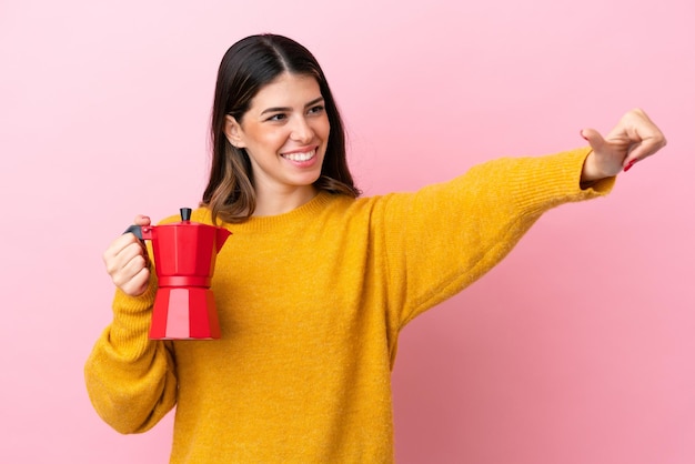 Jonge Italiaanse vrouw die een koffiezetapparaat houdt dat op roze achtergrond wordt geïsoleerd en een duim omhoog gebaar geeft