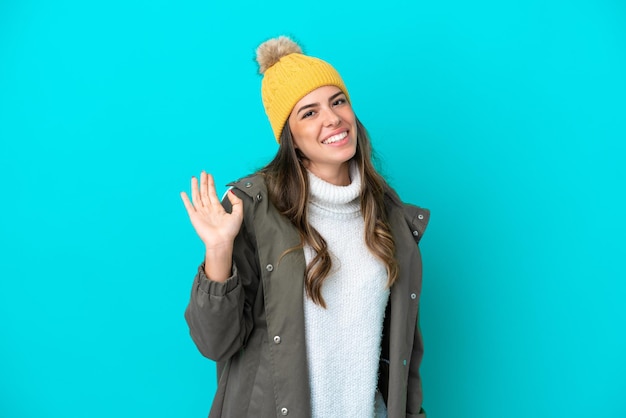 Jonge Italiaanse vrouw met een winterjas en muts die op een blauwe achtergrond wordt geïsoleerd en met de hand salueert met een blije uitdrukking