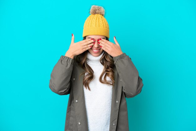 Jonge Italiaanse vrouw met een winterjas en muts geïsoleerd op een blauwe achtergrond die de ogen bedekt met de handen