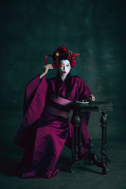 Jonge Japanse vrouw als geisha op donkergroene achtergrond. Retro stijl, vergelijking van tijdperken concept.