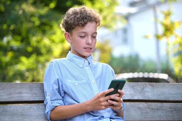 Jonge jongen die een spel speelt op zijn smartphone buiten in het zomerpark verslaving aan het concept van elektronische gadgets