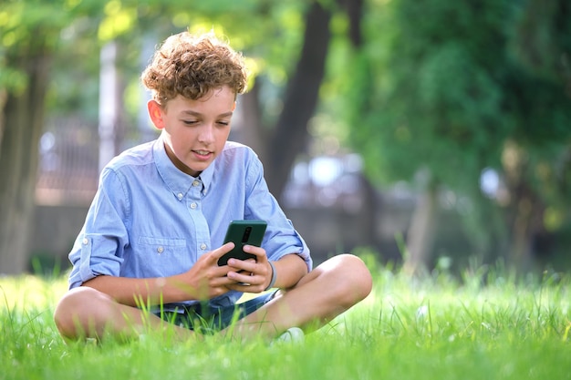 Jonge jongen die een spel speelt op zijn smartphone buiten in het zomerpark Verslaving aan het concept van elektronische gadgets
