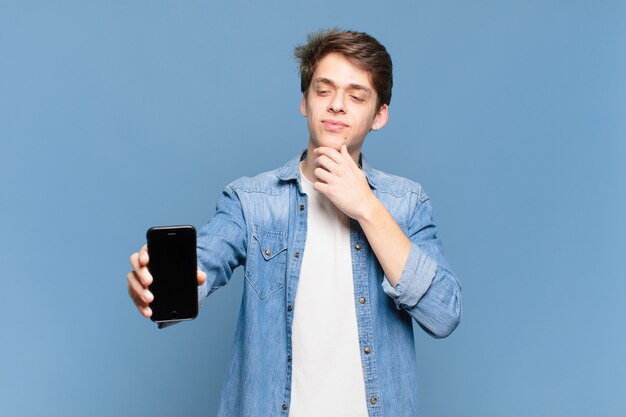 Jonge jongen glimlachend met een gelukkige, zelfverzekerde uitdrukking met de hand op de kin, zich afvragend en opzij kijkend. telefoon concept