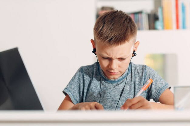 Jonge jongen in hoofdtelefoon zittend aan tafel met laptop en de voorbereiding op school thuis