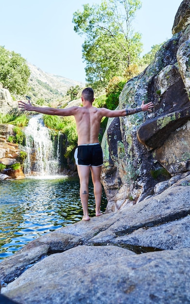 Jonge jongen met gekruiste armen achter zijn rug op een rots kijkend naar de rivier met waterval.