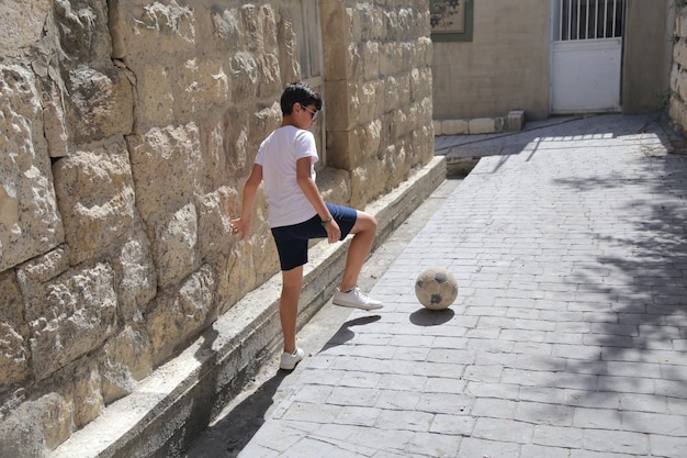 Jonge jongen spelen met een voetbal in een traditionele straat op de oude zomerachtergrond van het dorp