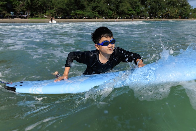 Foto jonge jongen student in surfen houdt softboard vast en probeert het terug in zee te brengen om te oefenen terwijl hij speelt tegen golven en opspattend water