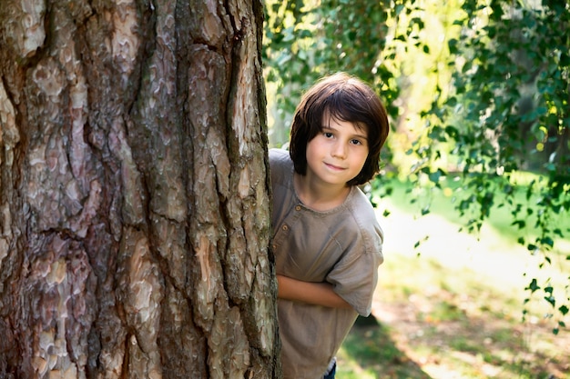 Jonge jongen van tien jaar oud verstopt achter een grote boom, glurend van achter een dikke stam. Portret van een kind in een park glimlachend en kijkend naar de camera.