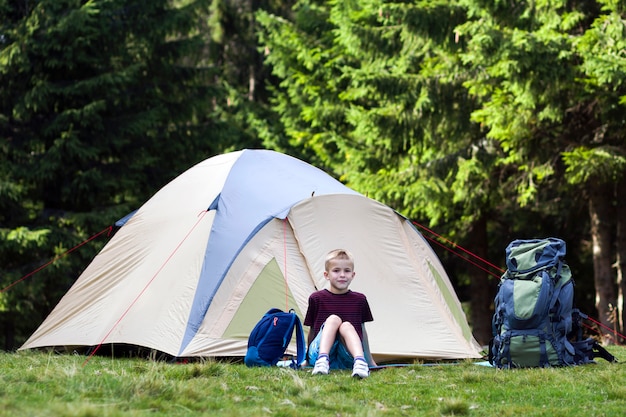 Jonge jongenszitting voor een tent dichtbij rugzakken die rust nemen