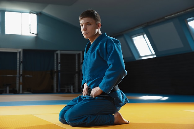 Jonge judo-kaukasische vechter in blauwe kimono met zwarte gordel die zelfverzekerd in de sportschool staat, sterk en gezond. Vechtvaardigheden in vechtsporten oefenen. Overwinnen, doel bereiken, zelfopbouw.