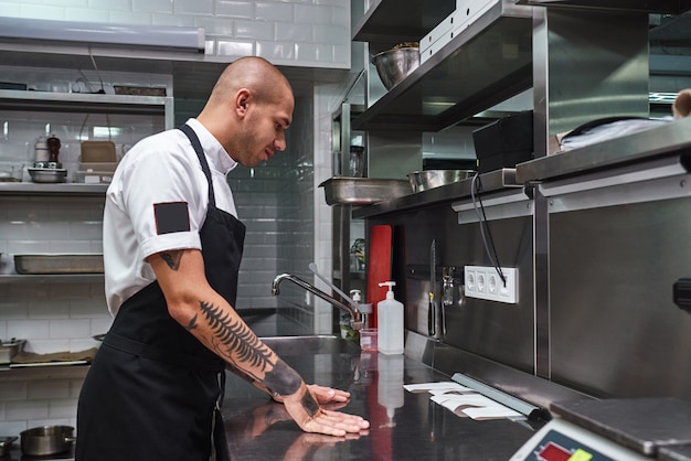 jonge kale mannelijke chef-kok met tatoeages die bestellijsten bekijkt op een stalen tafel in de keuken van een restaurant