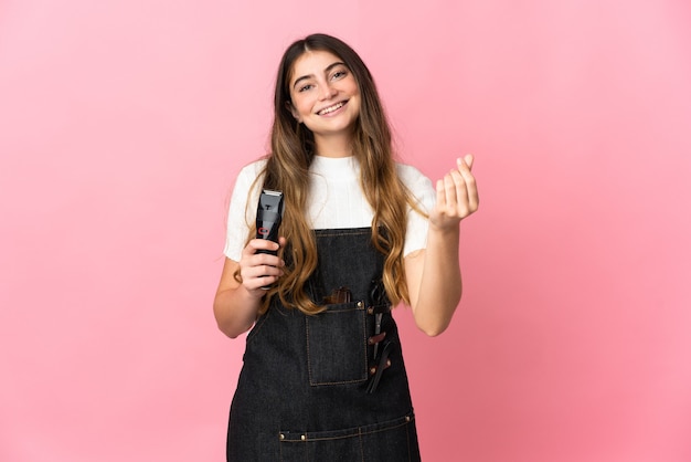 Jonge kappervrouw die op roze muur wordt geïsoleerd die geldgebaar maakt