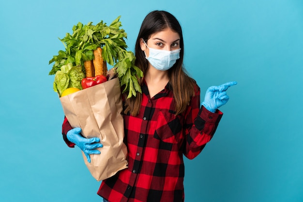 Jonge kaukasisch met groenten en masker geïsoleerd op blauw verrast en wijzende vinger naar de zijkant