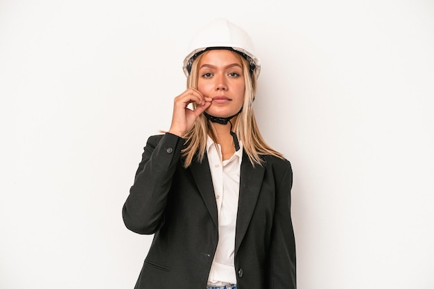 Jonge kaukasische architectenvrouw die een helm draagt die op witte achtergrond met vingers op lippen wordt geïsoleerd die een geheim houden.
