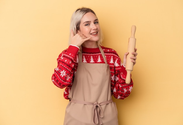 Jonge kaukasische bakkersvrouw die koekjes voor Kerstmis doet die op gele achtergrond worden geïsoleerd die een mobiel telefoongesprekgebaar met vingers tonen.