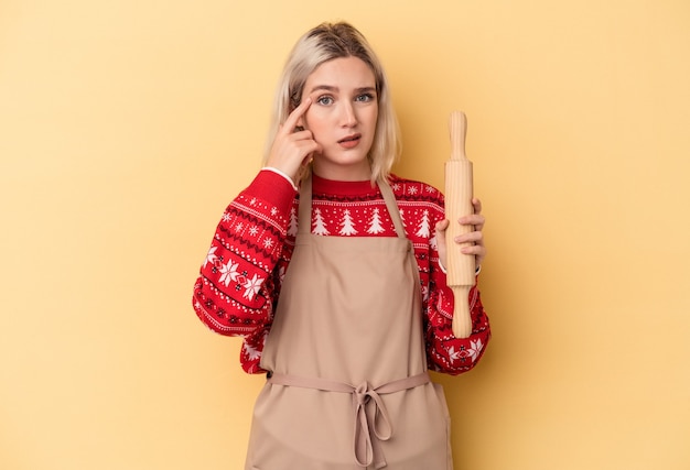 Jonge kaukasische bakkersvrouw die koekjes voor Kerstmis doet die op gele achtergrond worden geïsoleerd die een teleurstellingsgebaar met wijsvinger tonen.