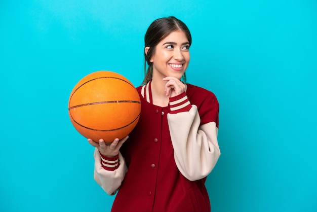 Jonge kaukasische basketbalspelervrouw die op blauwe achtergrond wordt geïsoleerd die aan de kant kijkt en glimlacht