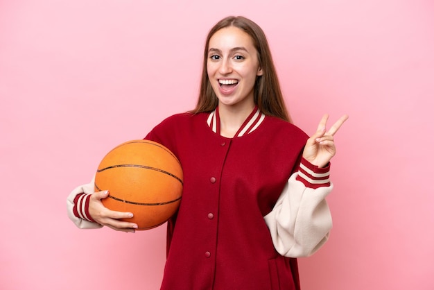 Jonge kaukasische basketbalspelervrouw over geïsoleerd op roze achtergrond die en overwinningsteken glimlacht toont