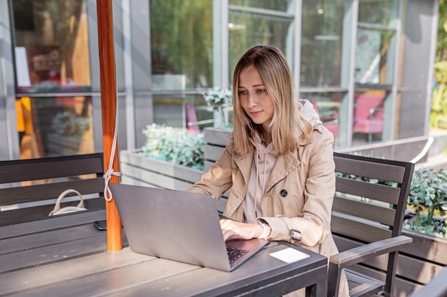 Jonge Kaukasische bedrijfsvrouw met blond haar die aan laptop in openluchtkoffie werkt.