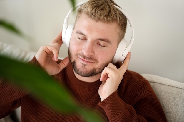 Jonge Kaukasische blond krullend haar man luisteren naar muziek met grote witte handsfree koptelefoon zittend op de Bank thuis op neutrale witte achtergrond knappe volwassen kerel portret lifestyleCopyspace